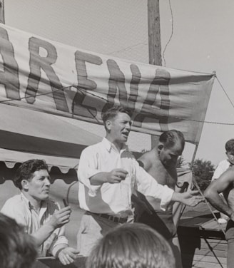 A group of men, some shirtless, are gathered around a platform or stage. A man in a white shirt appears to be speaking or gesturing to a crowd. A large sign with the word 'ARENA' is prominently displayed in the background. The scene seems to be set outdoors on a sunny day.