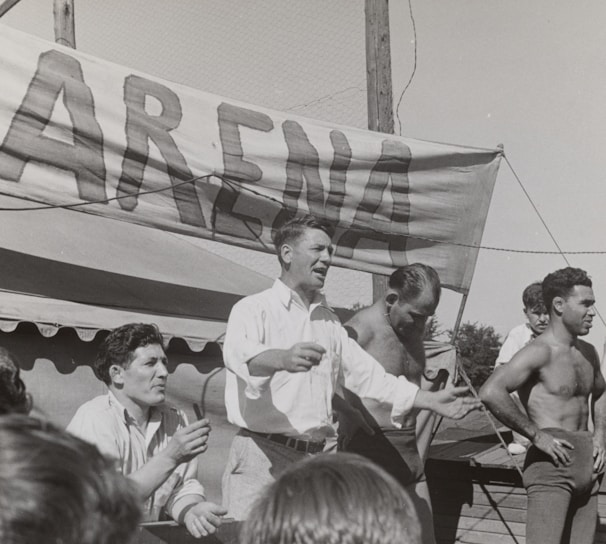 A group of men, some shirtless, are gathered around a platform or stage. A man in a white shirt appears to be speaking or gesturing to a crowd. A large sign with the word 'ARENA' is prominently displayed in the background. The scene seems to be set outdoors on a sunny day.
