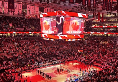 An indoor sports arena filled with spectators and a basketball court illuminated by red lighting. The scoreboards and video screens display promotional content, with numerous banners and championship flags hanging from the ceiling. Players and officials are gathered on the court, possibly in preparation for a game or event.