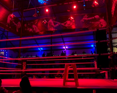 An empty boxing ring is illuminated by dramatic red and blue spotlights inside a dimly-lit arena. A small stool is placed in the center of the ring, surrounded by ropes. The arena is adorned with banners depicting action scenes, and a few silhouettes of spectators can be seen in the background.